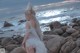 A woman in a white dress standing on a rocky beach.