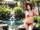 A woman in a red bikini sitting on a rock by a pool.