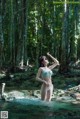 A woman in a bikini standing in a stream in the woods.