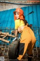 A woman in an orange hard hat and overalls leaning on a tire.