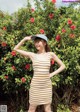 A woman in a striped dress and hat posing in front of a bush.