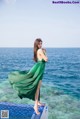 A woman in a green dress standing on a ledge by the ocean.